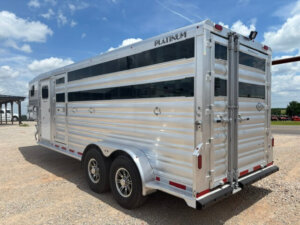 A shiny silver Platinum stock combo livestock trailer, 20' long with black-tinted windows and dual axles, is parked on a gravel lot under a partly cloudy sky. -Stillwater Trailer Sales