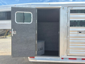 The image shows an open door of a 20' Platinum Stock Combo horse trailer, revealing a carpeted tack room inside with a small bench or storage compartment against the back wall. The exterior is metallic with horizontal panels. -Stillwater Trailer Sales