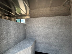 The interior of a Platinum Stock Combo trailer with gray carpeted walls and partitions, a small window, and an angled bench or platform under the window; metallic ceiling beams are visible above in this spacious 20' model. -Stillwater Trailer Sales