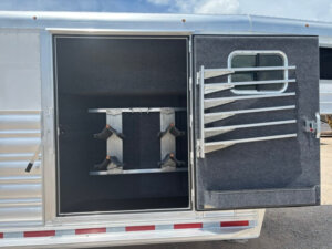 Open compartment of a 20' Platinum Stock Combo horse trailer with the door swung wide, showing interior saddle racks and metal bars on the window. The silver trailer has a carpeted interior and red-and-white safety stripes. -Stillwater Trailer Sales