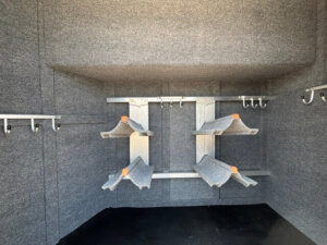 Gray, carpeted interior of a Platinum Stock Combo storage shed featuring angled racks with felt padding and multiple metal hooks on the walls, designed for organizing and holding items securely in a spacious 20' layout. -Stillwater Trailer Sales