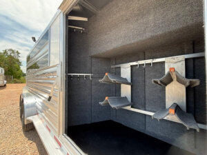 The image shows the interior of an empty 20&rsquo; Platinum Stock Combo horse trailer with carpeted walls, saddle racks, and bridle hooks for organized tack storage. The trailer is open, with sunlight visible outside. -Stillwater Trailer Sales