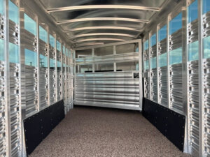 Interior of an empty Platinum Stock Combo 20' aluminum livestock trailer with ribbed metal walls, a textured non-slip floor, and a dividing gate at the front. Light streams in through narrow side vents and ceiling panels. -Stillwater Trailer Sales