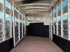 Interior view of a 20' Platinum Stock Combo trailer, featuring metal slatted walls, a textured non-slip floor, and a partition dividing the space. The curved roof has support beams with natural light streaming through the slats. -Stillwater Trailer Sales