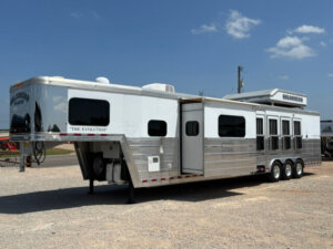 A large silver and white horse trailer labeled The Evolution sits parked on gravel under a partly cloudy sky. The trailer has multiple windows, vents, and a slide-out section on the side. -Stillwater Trailer Sales