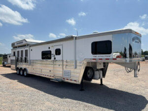 A large, white and silver multi-axle gooseneck horse trailer named The Evolution is parked on gravel under a partly cloudy sky. The trailer has multiple windows and a shiny exterior finish. -Stillwater Trailer Sales