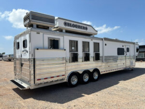 A large silver and white Bloomer horse trailer with three axles is parked on a gravel lot under a blue sky, featuring multiple windows and an air conditioning unit on the roof. -Stillwater Trailer Sales