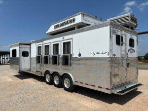 A large, silver and white Bloomer horse trailer with multiple windows and vents, parked on gravel under a partly cloudy sky. The trailer has the slogan only the best written on the side. -Stillwater Trailer Sales