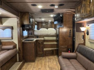 Interior of an RV with wood-paneled walls, brown leather sofas, a small kitchen area with a sink, microwave, and cabinets, stairs leading to a lofted bed, and a mounted TV on the right wall. -Stillwater Trailer Sales