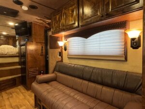 A cozy RV interior featuring a brown leather sofa, matching wood cabinets, a window with blinds, wall-mounted lights, and a visible sleeping area with bedding in the background. -Stillwater Trailer Sales