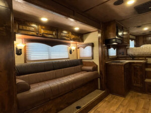 A cozy RV interior with a brown leather couch, wooden cabinets, wall lights, and a small kitchen area featuring a sink and stove. Steps lead up to a raised bed area with additional storage and window blinds. -Stillwater Trailer Sales