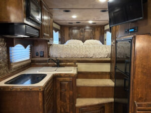 Interior of an RV with a compact kitchen, including a sink, stovetop, microwave, and cabinets. Carpeted steps lead up to a bed with a white comforter, and a flat-screen TV is mounted near a refrigerator. -Stillwater Trailer Sales