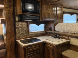 A compact RV kitchen features dark wood cabinets, a built-in microwave, a stove, a sink, and decorative backsplash. There&rsquo;s a window with a scalloped valance and a cozy sleeping area with a textured cream comforter above. -Stillwater Trailer Sales