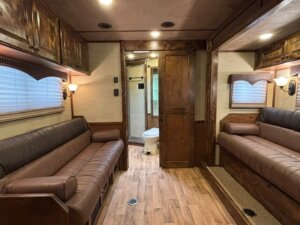 Interior of a trailer with wood paneling, two brown leather sofas facing each other, wall lamps, overhead cabinets, and a view into a bathroom with a toilet at the back. -Stillwater Trailer Sales
