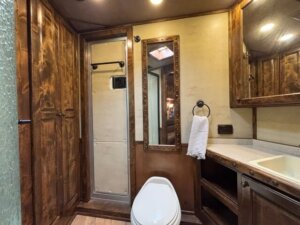 A rustic-style bathroom with wooden cabinets, a toilet, a tall mirror, a countertop with a sink, an open shelf, a towel ring holding a white towel, and a metal door with a towel bar. -Stillwater Trailer Sales