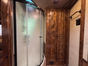 A bathroom with a curved glass shower enclosure and a tall wooden cabinet with two doors, alongside a beige wall with a towel bar. -Stillwater Trailer Sales