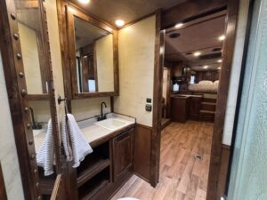 A view from a bathroom with a sink, mirror, striped towel, and wood accents, looking into a cozy RV interior with wood flooring and a bed in the background. -Stillwater Trailer Sales