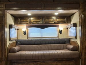 A brown leather couch is set against a wall with three windows covered by blinds, surrounded by wooden cabinets and two wall sconces in a cozy, wood-paneled room. -Stillwater Trailer Sales