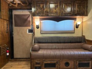 A cozy RV interior features a brown leather couch, wooden cabinets, wall-mounted lamps, a shaded window, and a door with a fire extinguisher nearby. The decor is rustic with earthy tones. -Stillwater Trailer Sales