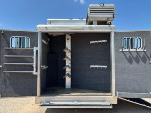 Open trailer compartment with carpeted interior, multiple hooks and racks on the walls and doors, and saddle stands in the center, designed for organizing equestrian or tack equipment under a clear blue sky. -Stillwater Trailer Sales