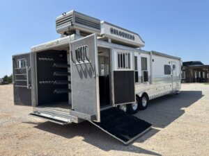 A large white horse trailer labeled BLOOMER with its rear doors open, revealing interior storage racks and a black loading ramp extended onto a gravel lot under a clear blue sky. -Stillwater Trailer Sales