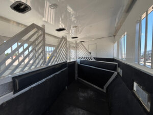 Interior of a horse trailer with metal dividers, padded walls, barred windows, and black flooring, designed to safely transport multiple horses. Sunlight shines through the windows, illuminating the clean space. -Stillwater Trailer Sales