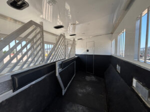 Interior of an empty livestock or horse trailer with padded dividers, slatted metal partitions, black walls, and windows letting in daylight. The floor appears worn, and the trailer door is visible at the far end. -Stillwater Trailer Sales