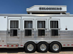 A silver Bloomer horse trailer with three sets of barred windows and three tandem wheels, parked outdoors under a clear blue sky. -Stillwater Trailer Sales